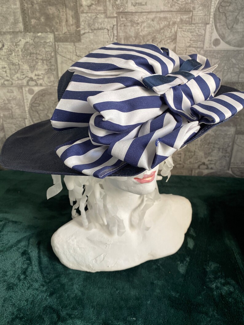 May include: A navy blue sun hat with a wide brim and a large bow made of white and navy blue striped fabric. The hat is displayed on a white bust, highlighting its design and style.