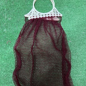 Peut inclure: Un sac en filet bordeaux avec une poignée en plastique blanc. La poignée est circulaire et fixée à un dessus décoratif en plastique blanc. Le sac est fait d'un matériau en filet rouge foncé.