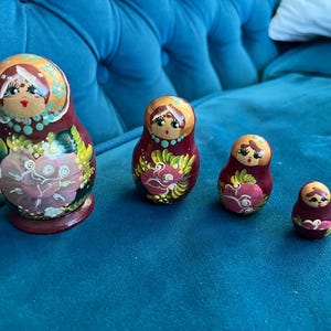 Op de afbeelding: Set van vier traditionele Russische nestpoppen, ook wel matroesjka-poppen genoemd. De poppen zijn geschilderd in een levendig rood, roze en groen kleurenschema met bloemmotieven. De grootste pop is ongeveer 7,5 cm hoog.