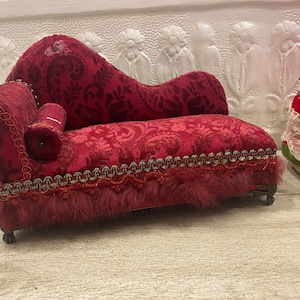 May include: A small red velvet chaise longue with a tufted back and a decorative trim. The chaise has a small pillow and is on a white surface.