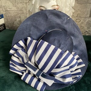 May include: A navy blue sun hat with a wide brim and a striped bow. The bow features alternating white and navy blue stripes. The hat is displayed on a mannequin head.