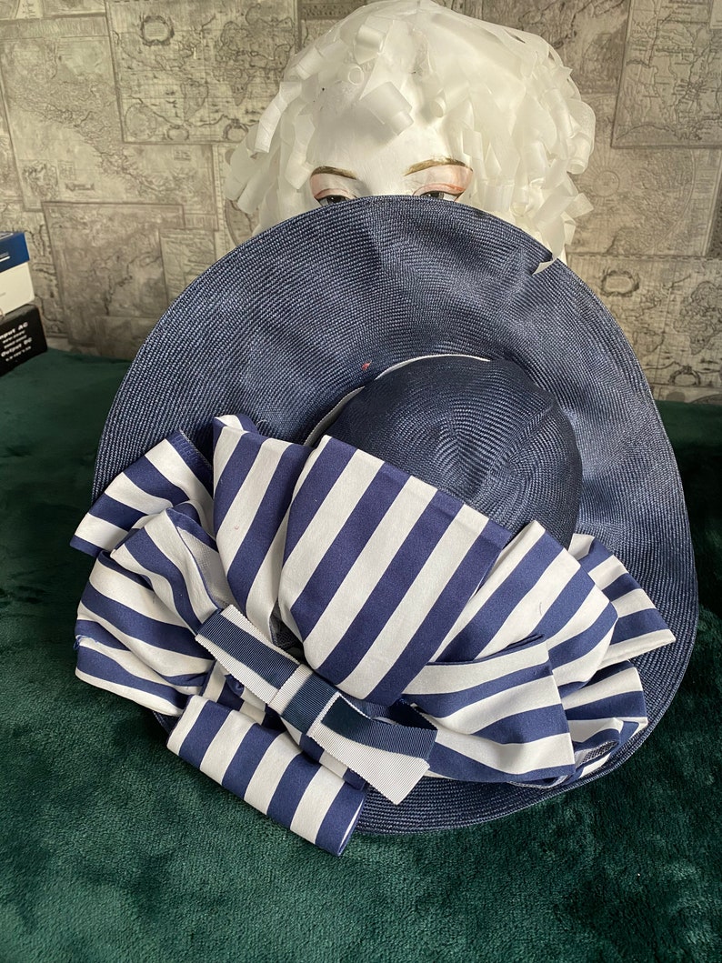 May include: A navy blue sun hat with a wide brim and a striped bow. The bow features alternating white and navy blue stripes. The hat is displayed on a mannequin head.