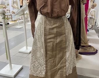 May include: A vintage brown dress with a long-sleeved top and a long skirt. The skirt has a cream-coloured lace panel on the front. The dress is displayed on a mannequin in a museum, showcasing historical fashion.