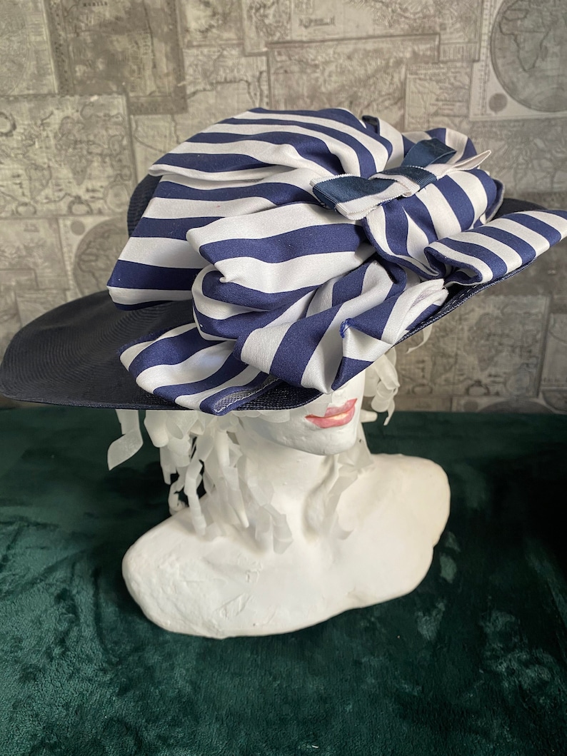 May include: A navy blue and white striped sun hat with a wide brim. The hat features a large, decorative bow made of the same striped fabric. The hat is displayed on a white bust.
