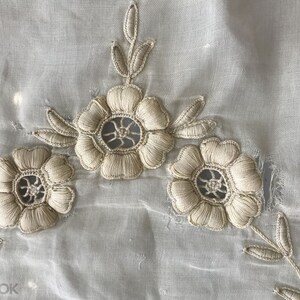 May include: White sheer fabric with delicate embroidered flowers. The flowers are beige and have a lacy design.