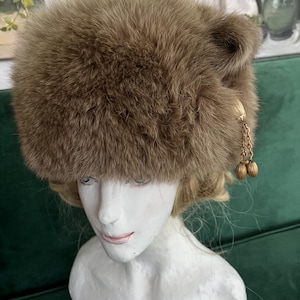 Puede incluir: Un sombrero de piel sintética marrón con una pequeña solapa de oreja en un lado. El sombrero tiene una cadena dorada con dos abalorios redondos dorados que cuelgan.