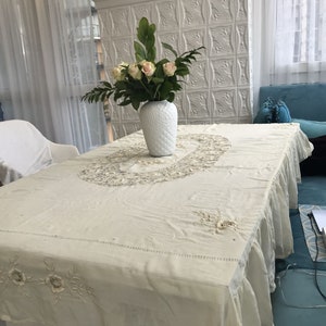 May include: A white tablecloth with delicate embroidery and a ruffled edge. A white vase with a floral design holds a bouquet of pink roses and green leaves on a table.
