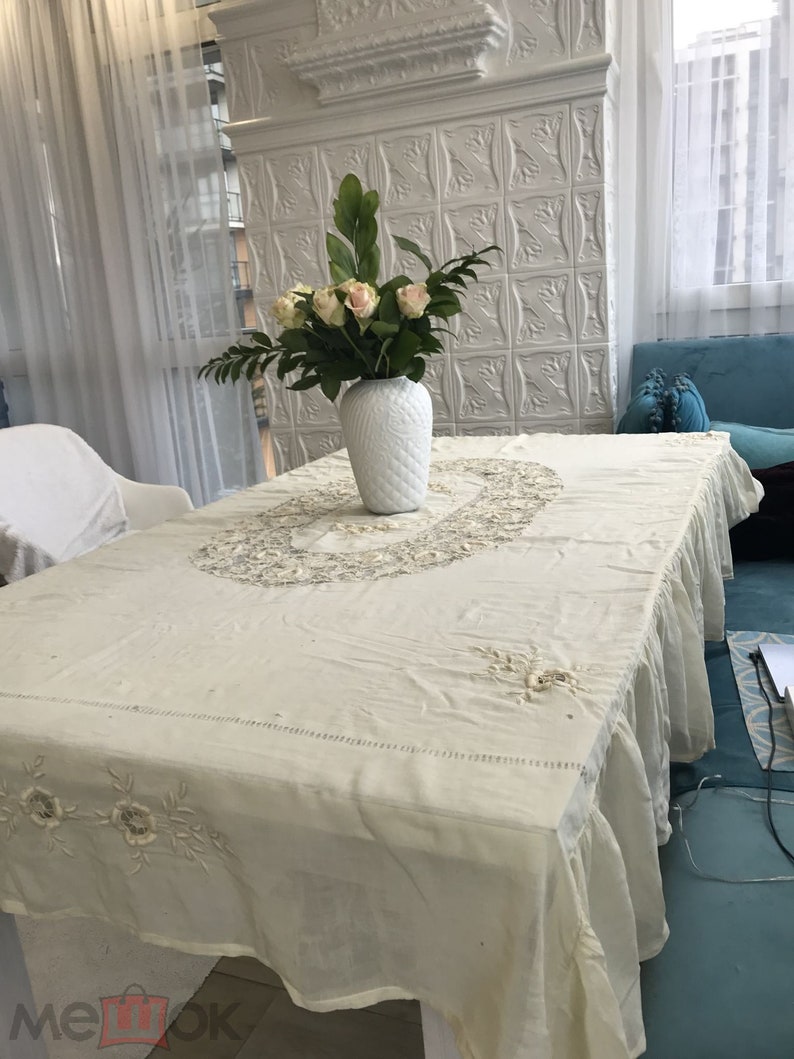 May include: A white tablecloth with delicate embroidery and a ruffled edge. A white vase with a floral design holds a bouquet of pink roses and green leaves on a table.
