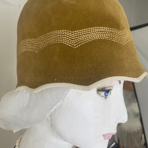 May include: A brown velvet cloche hat with a white stitched design around the crown. The hat has a white trim around the brim.