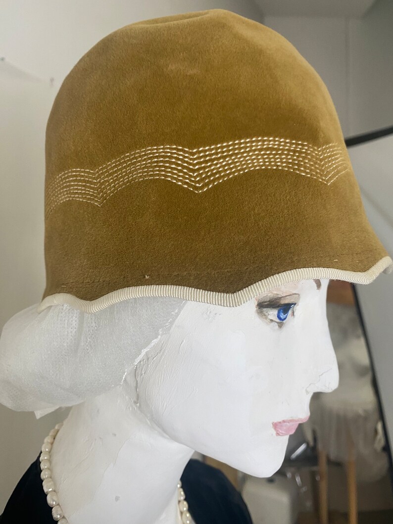 May include: A brown velvet cloche hat with a white stitched design around the crown. The hat has a white trim around the brim.