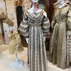 Puede incluir: Tres maniquíes exhiben vestidos vintage. El maniquí central viste un vestido a rayas marrón y crema con mangas abullonadas. Un maniquí más pequeño a la izquierda lleva un vestido amarillo. El maniquí de la derecha lleva un vestido dorado.