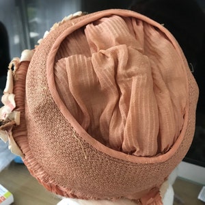 May include: A light brown hat with a ruffled trim and a soft, peach-colored fabric lining. The hat is made of a woven material and has a slightly textured surface.