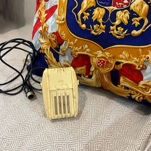 May include: A vintage, off-white microphone with a black cord and connector. The microphone is rectangular with vertical slots. A decorative pillow with a crest design in gold, red, and blue is in the background.