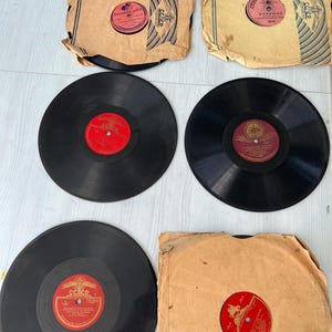 Może przedstawiać: Kolekcja zabytkowych płyt winylowych. Czarne płyty mają czerwone etykiety z tekstem. Niektóre płyty znajdują się w postarzanych papierowych kopertach z nadrukami i etykietami. Płyty są ułożone na jasnej powierzchni.