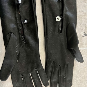 May include: A pair of black gloves with a button closure. The gloves are long, extending past the wrist, and feature small, decorative buttons. The fabric appears to be a smooth, matte material.