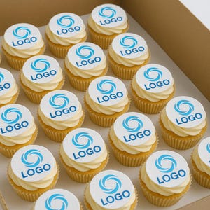 Op de afbeelding: Een doosje cupcakes met witte frosting en eetbare logo-toppers. De cupcakes hebben een gele basis en zijn in rijen gerangschikt. Het logo is een blauw wervelontwerp met het woord "LOGO".