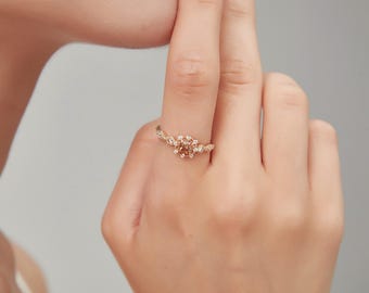 May include: A gold-toned ring with a central, round, light brown gemstone. The ring features a floral design around the center stone and smaller accent stones along the band. The ring is on a person's finger.