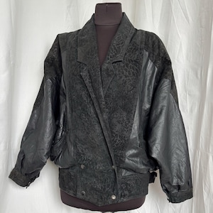 May include: Black leather jacket with a textured pattern on the upper chest and shoulders. The jacket has a double-breasted design with two rows of buttons and a wide lapel. The sleeves are a smooth leather material. The jacket is displayed on a mannequin.