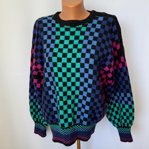 May include: A colorful, vintage sweater with a checkerboard pattern. The sweater features a black base with squares of teal, blue, purple, and pink. The cuffs and hem have a striped design. The sweater is displayed on a mannequin.