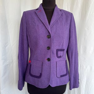 May include: A purple blazer with a two-button closure, featuring two front pockets and a notched lapel. The blazer has dark purple trim on the pockets and a red accent on the lapel. The jacket is displayed on a black mannequin.