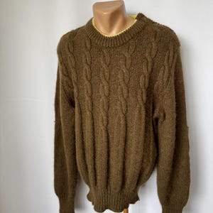 Könnte beinhalten: Ein brauner Zopfstrickpullover mit gelbem Rundhalsausschnitt. Der Pullover hat lange Ärmel und einen gerippten Saum. Der Pullover wird auf einer Schaufensterpuppe vor weißem Hintergrund präsentiert.