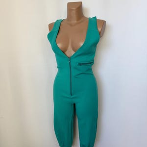 Op de afbeelding: Tealkleurige jumpsuit met een geribbelde textuur. Het mouwloze kledingstuk heeft een diepe V-hals, een ritssluiting aan de voorkant en een klein borstzakje. De pijpen lopen taps toe naar geribbelde manchetten. De mannequin is ongeveer 152 cm lang.