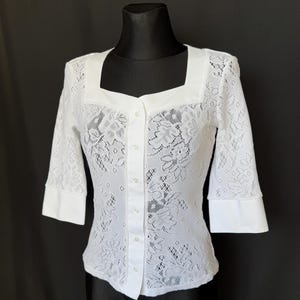 May include: A white lace blouse featuring a square neckline, three-quarter length sleeves with solid white cuffs, and a front button-down closure. The garment is constructed from a floral patterned lace fabric with a solid white placket.