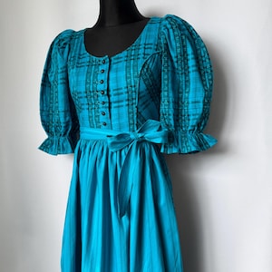 Puede incluir: Un vestido turquesa con corpi&ntilde;o a cuadros y falda lisa. El corpi&ntilde;o tiene botones y mangas abullonadas. Una faja a juego se ata a la cintura. El vestido se exhibe en un maniqu&iacute; negro.