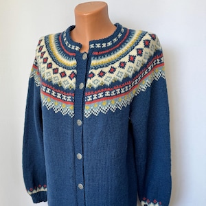 Könnte beinhalten: Ein blauer Strickcardigan mit einem bunten geometrischen Muster an Joch und Bündchen. Der Cardigan hat einen Knopfverschluss und lange Ärmel.