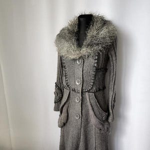 Puede incluir: Un abrigo de lana gris con cuello de piel sintética. El abrigo tiene una botonadura frontal, bolsillos y costuras decorativas. El abrigo largo se exhibe en un maniquí.