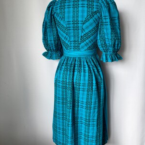 Puede incluir: Un vestido turquesa vintage con un estampado de cuadros. El vestido presenta mangas cortas abullonadas con pu&ntilde;os con volantes, un corpi&ntilde;o entallado y una falda fruncida. Un cintur&oacute;n a juego ci&ntilde;e la cintura. El vestido se exhibe en un maniqu&iacute;.