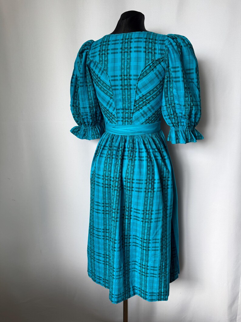 Puede incluir: Un vestido turquesa vintage con un estampado de cuadros. El vestido presenta mangas cortas abullonadas con pu&ntilde;os con volantes, un corpi&ntilde;o entallado y una falda fruncida. Un cintur&oacute;n a juego ci&ntilde;e la cintura. El vestido se exhibe en un maniqu&iacute;.