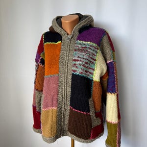 Può includere: Una giacca con cappuccio e cerniera realizzata con vari inserti in maglia nei colori arancione, viola, nero, marrone, rosa e crema. Gli inserti sono uniti da cuciture a punto croce visibili con fili dai colori neon e pastello a contrasto.