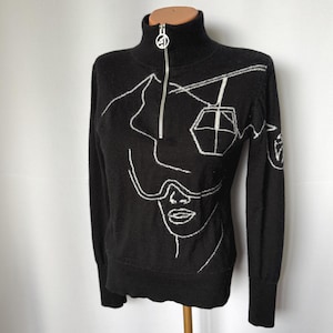 May include: Black turtleneck sweater featuring a half-zip and a white embroidered ski design. The design includes a face, ski goggles, and a ski lift. The sweater has long sleeves and ribbed cuffs.