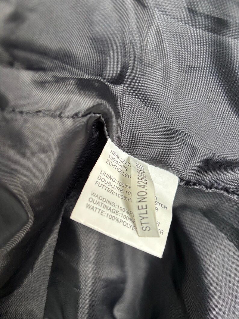 May include: Close-up of a black garment's interior, revealing a tag with text in multiple languages. The tag lists the materials: real leather, lining, wadding, and other components. The style number is 42500.