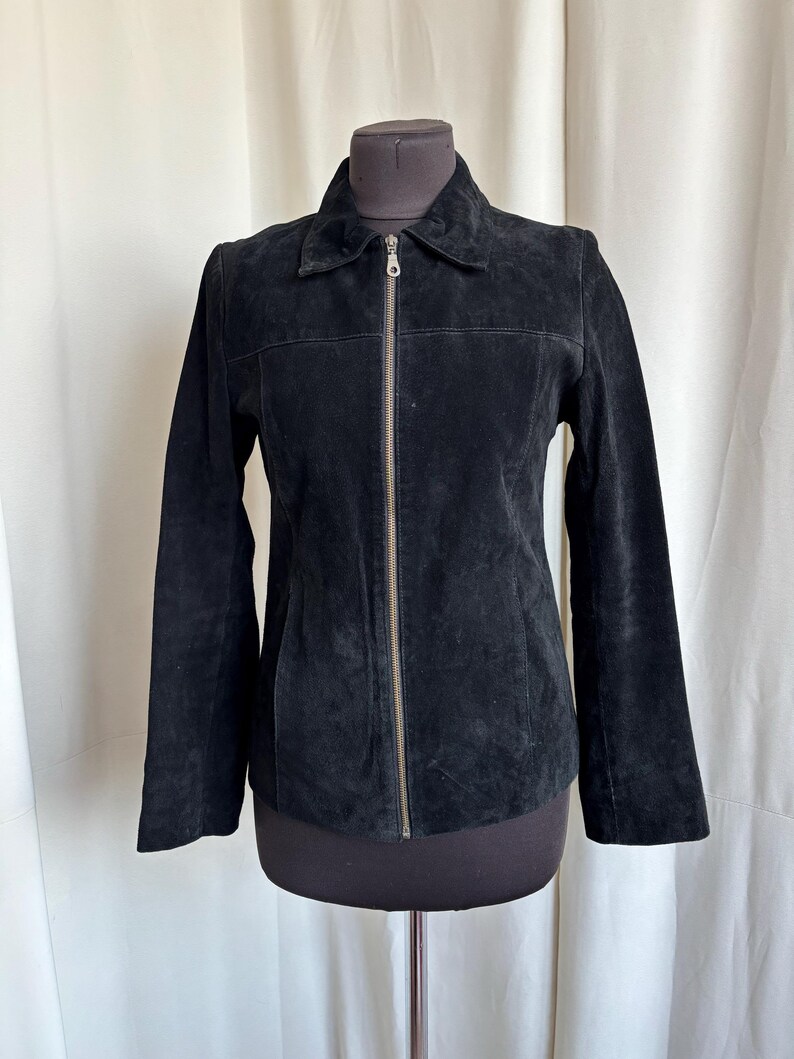 May include: A black suede jacket with a collared neckline and a full-length zipper closure. The jacket is displayed on a mannequin and has long sleeves. The suede material has a soft, textured appearance.