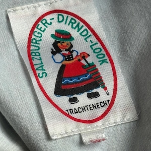 Puede incluir: Una etiqueta blanca con el texto "SALZBURGER-DIRNDL-LOOK" en un &oacute;valo rojo. La etiqueta presenta una caricatura de una mujer con ropa tradicional austriaca, sosteniendo una aguja de tejer. El texto "TRACHTENECHT" est&aacute; debajo.