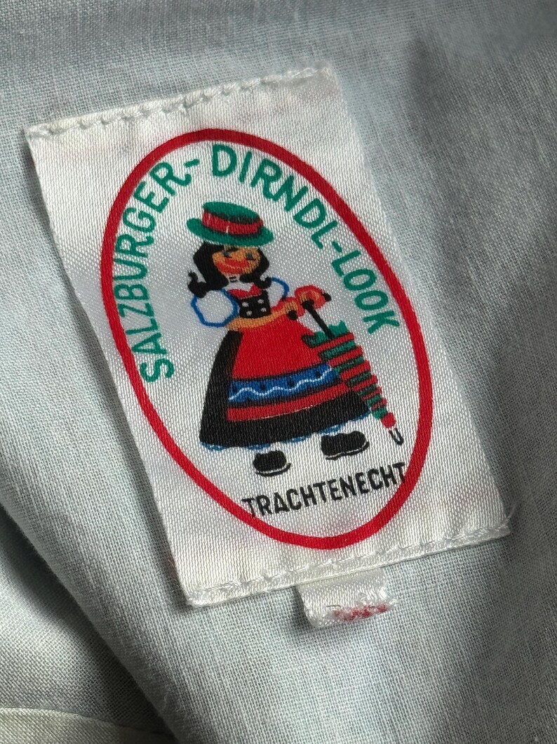 Puede incluir: Una etiqueta blanca con el texto "SALZBURGER-DIRNDL-LOOK" en un &oacute;valo rojo. La etiqueta presenta una caricatura de una mujer con ropa tradicional austriaca, sosteniendo una aguja de tejer. El texto "TRACHTENECHT" est&aacute; debajo.
