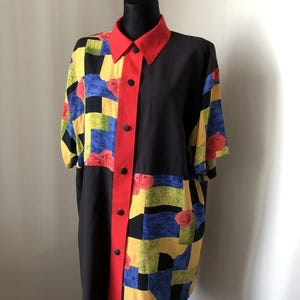 Puede incluir: Camisa vintage en maniquí. La camisa tiene cuerpo negro con cuello y tapeta rojos. Las mangas y la parte superior presentan un colorido diseño patchwork con motivos florales amarillos, azules y rojos. Camisa de manga corta.