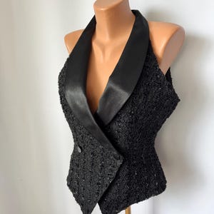 Puede incluir: Un chaleco negro sin mangas con cuello de chal de satén y cierre frontal cruzado con dos botones. El cuerpo principal de la prenda está confeccionado con un tejido de encaje negro texturizado e intrincado con sutiles detalles de lentejuelas.