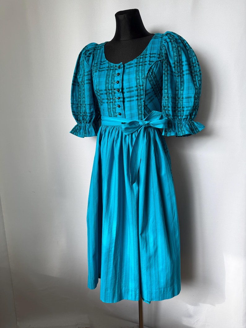 Puede incluir: Un vestido turquesa con corpi&ntilde;o a cuadros y falda lisa. El corpi&ntilde;o tiene botones y mangas abullonadas. Una faja a juego se ata a la cintura. El vestido se exhibe en un maniqu&iacute; negro.