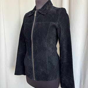 May include: Black suede jacket with a collar and a full-length zipper. The jacket has long sleeves and is displayed on a mannequin. The suede appears soft and has a matte finish. The jacket is a classic style.