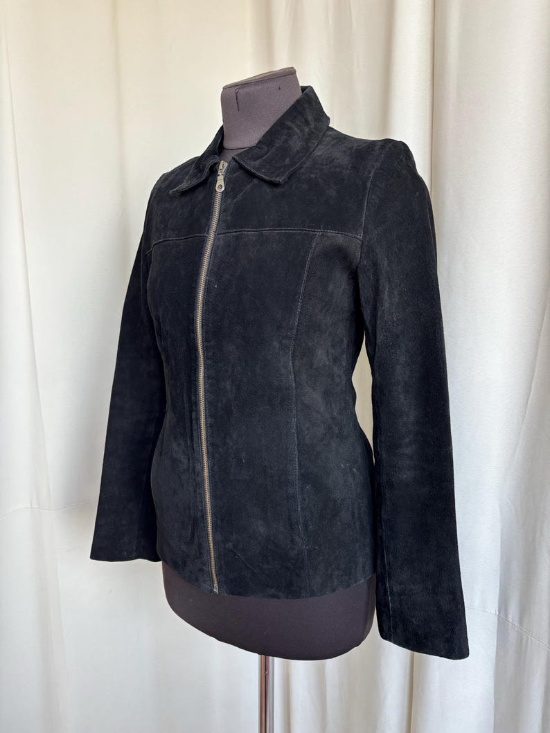 May include: Black suede jacket with a collar and a full-length zipper. The jacket has long sleeves and is displayed on a mannequin. The suede appears soft and has a matte finish. The jacket is a classic style.