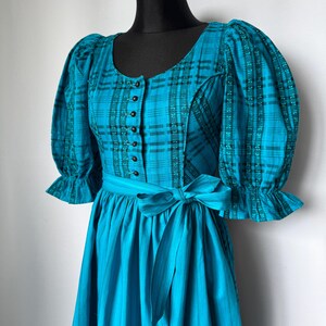 Puede incluir: Un vestido a cuadros turquesa y negro con un corpi&ntilde;o ajustado y una falda amplia. El vestido presenta un escote redondo, mangas abullonadas y un cintur&oacute;n a juego. El corpi&ntilde;o tiene una fila de botones negros.