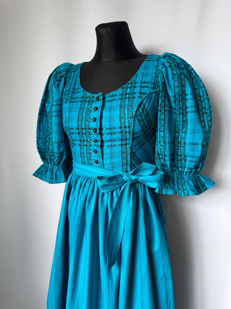 Puede incluir: Un vestido a cuadros turquesa y negro con un corpi&ntilde;o ajustado y una falda amplia. El vestido presenta un escote redondo, mangas abullonadas y un cintur&oacute;n a juego. El corpi&ntilde;o tiene una fila de botones negros.