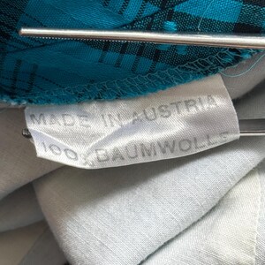 Puede incluir: Primer plano de una etiqueta que dice "MADE IN AUSTRIA" y "100% BAUMWOLLE" sobre una tela azul claro. La tela tiene un patr&oacute;n de cuadros turquesa y negro. Tambi&eacute;n se ve un objeto met&aacute;lico plateado.