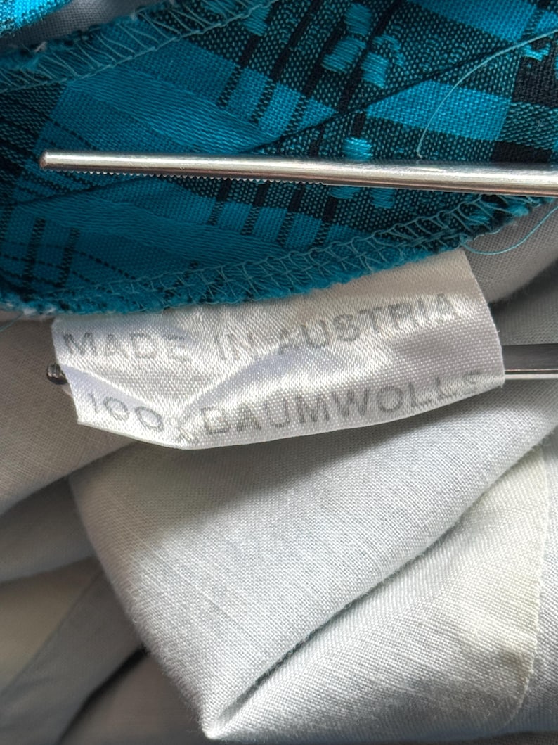 Puede incluir: Primer plano de una etiqueta que dice "MADE IN AUSTRIA" y "100% BAUMWOLLE" sobre una tela azul claro. La tela tiene un patr&oacute;n de cuadros turquesa y negro. Tambi&eacute;n se ve un objeto met&aacute;lico plateado.