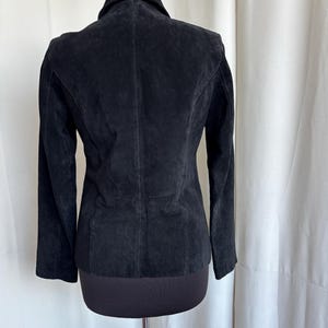 May include: A black suede jacket with a classic collar and long sleeves. The jacket is displayed on a mannequin, showcasing its tailored fit and the texture of the suede. The back view highlights the seams and design details.