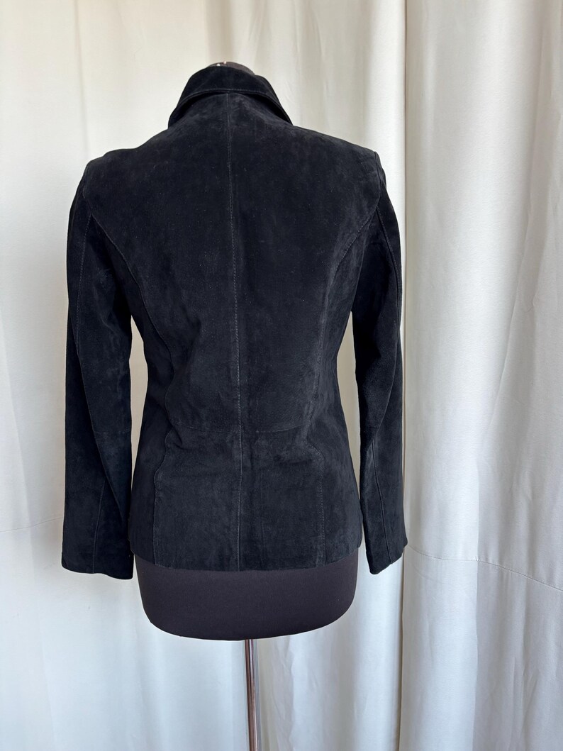 May include: A black suede jacket with a classic collar and long sleeves. The jacket is displayed on a mannequin, showcasing its tailored fit and the texture of the suede. The back view highlights the seams and design details.