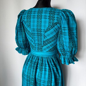 Puede incluir: Vestido vintage de color turquesa con estampado de cuadros, con mangas abullonadas y corpi&ntilde;o entallado. El vestido tiene una faja ancha turquesa en la cintura y pu&ntilde;os con volantes. El vestido se exhibe en un maniqu&iacute; negro.